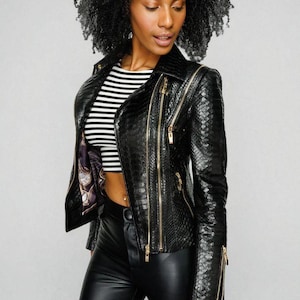 May include: A black faux leather jacket with a snakeskin texture and gold zippers. The jacket has a classic biker style with multiple zippers and a notched collar. The model is wearing a black and white striped top and black leather pants.