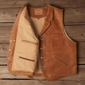 May include: A brown leather vest with a notched lapel and four silver buttons. The vest has two front pockets and a tan interior lining with a zippered pocket. The vest is open, revealing the interior construction and details.