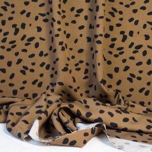 May include: Brown fabric with a black spot pattern. The fabric is draped, showing folds and texture. The pattern consists of irregular black spots scattered across the brown material. The fabric is likely used for clothing or home decor.