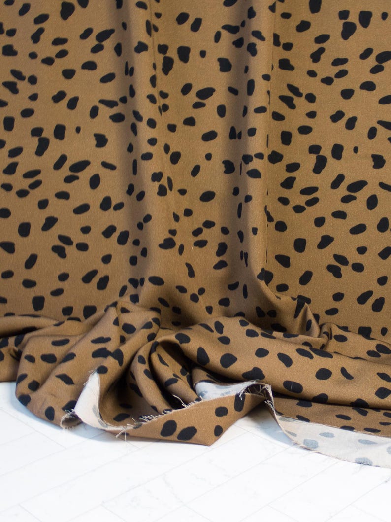 May include: Brown fabric with a black spot pattern. The fabric is draped, showing folds and texture. The pattern consists of irregular black spots scattered across the brown material. The fabric is likely used for clothing or home decor.