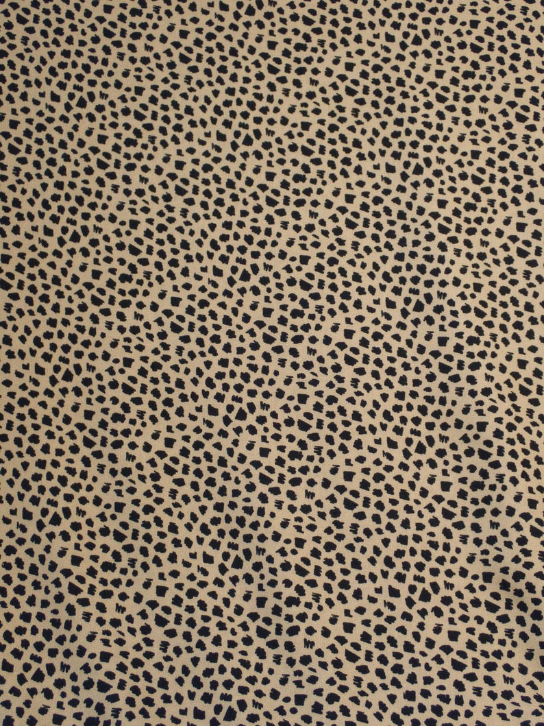 May include: A close-up of a fabric with a tan background and a repeating pattern of black, irregular shapes. The shapes are small and densely packed, creating a speckled appearance across the material.