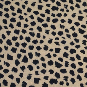 May include: Close-up of a fabric with a repeating pattern of dark navy blue irregular shapes on a tan background. The shapes vary in size and density, creating a spotted or speckled effect across the textile.