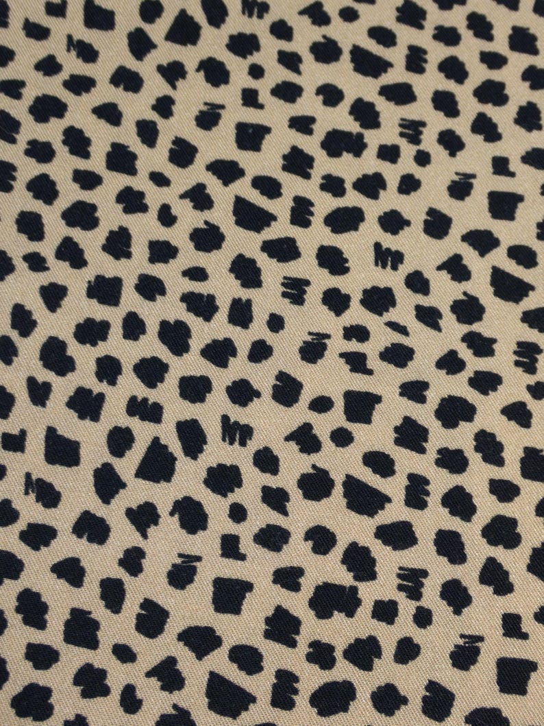 May include: Close-up of a fabric with a repeating pattern of dark navy blue irregular shapes on a tan background. The shapes vary in size and density, creating a spotted or speckled effect across the textile.