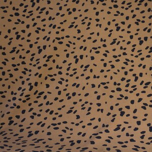 May include: A close-up of a fabric with a repeating pattern of black, irregular shapes on a tan background. The shapes are scattered across the surface, creating a spotted or speckled effect. The fabric appears to be smooth.