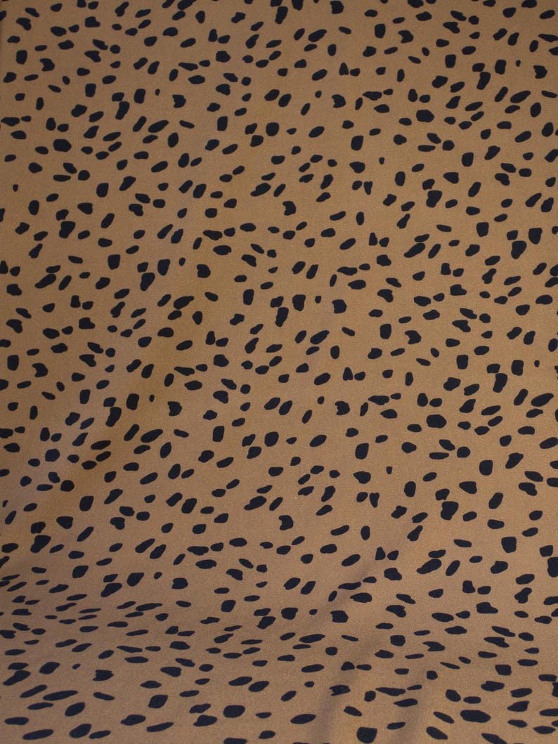 May include: A close-up of a fabric with a repeating pattern of black, irregular shapes on a tan background. The shapes are scattered across the surface, creating a spotted or speckled effect. The fabric appears to be smooth.