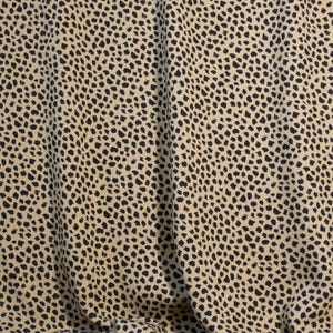 May include: Close-up of a fabric with a tan background and black spots. The fabric is draped, creating folds and shadows. The pattern is reminiscent of animal print.