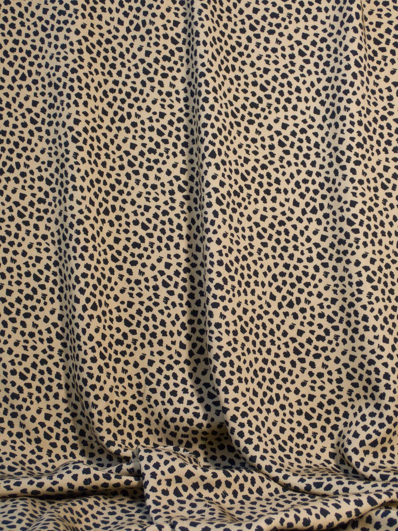 May include: Close-up of a fabric with a tan background and black spots. The fabric is draped, creating folds and shadows. The pattern is reminiscent of animal print.