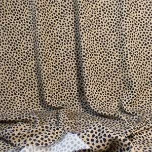 May include: Tan fabric with a black spotted pattern, reminiscent of a cheetah's coat. The fabric is draped, showing folds and the raw edge of the material. The pattern consists of irregular black spots on a tan background.