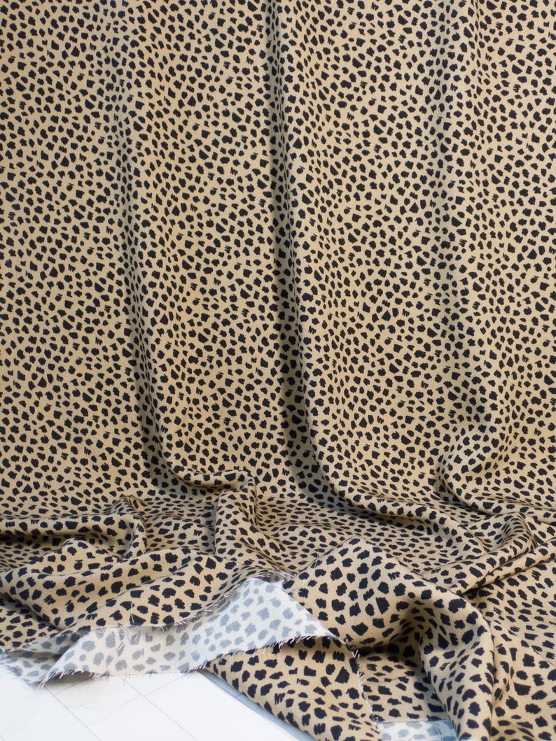 May include: Tan fabric with a black spotted pattern, reminiscent of a cheetah's coat. The fabric is draped, showing folds and the raw edge of the material. The pattern consists of irregular black spots on a tan background.