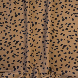 May include: Tan fabric with a repeating pattern of black, irregular spots. The fabric is draped, showing folds and shadows. The overall texture appears soft and possibly suitable for clothing or home decor.