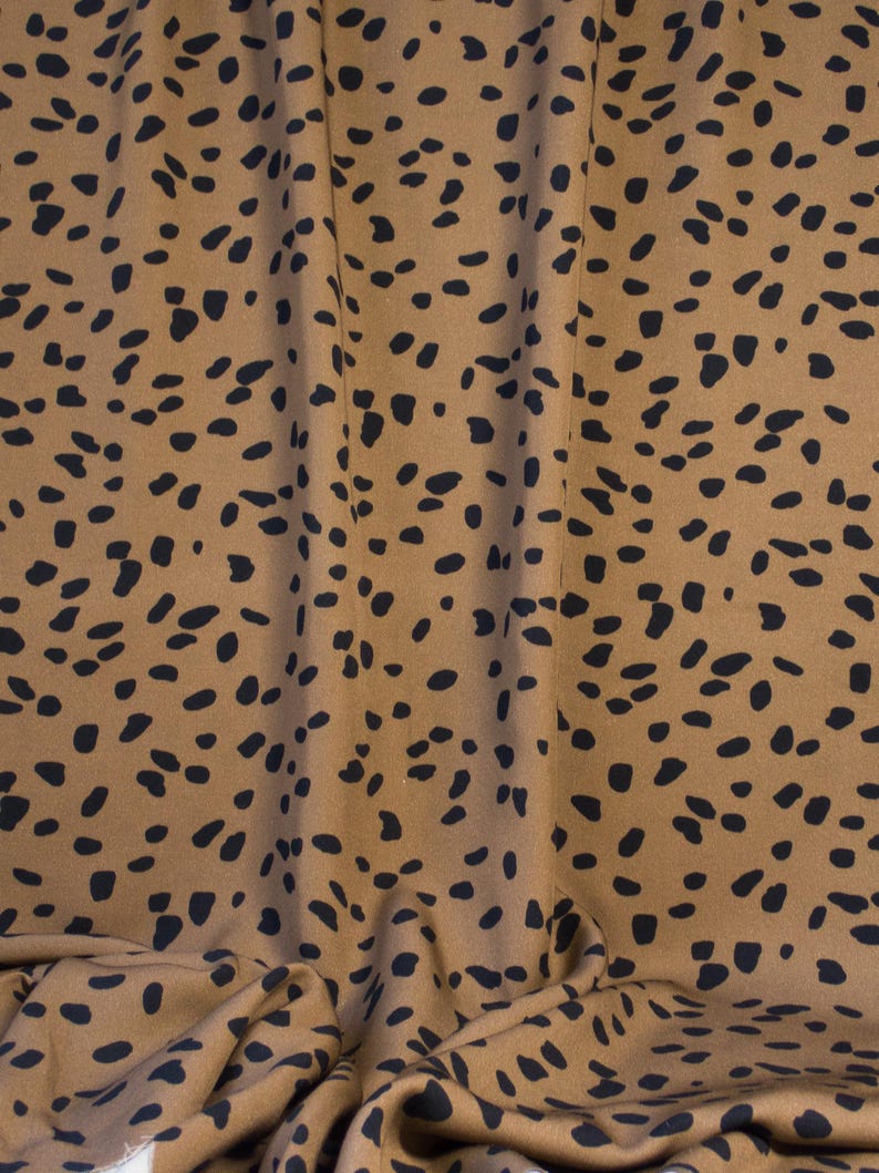 May include: Tan fabric with a repeating pattern of black, irregular spots. The fabric is draped, showing folds and shadows. The overall texture appears soft and possibly suitable for clothing or home decor.