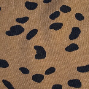 May include: Close-up of a tan fabric with a repeating pattern of dark navy blue irregular shapes. The shapes are scattered across the surface, creating a spotted or speckled effect. The fabric has a subtle texture.