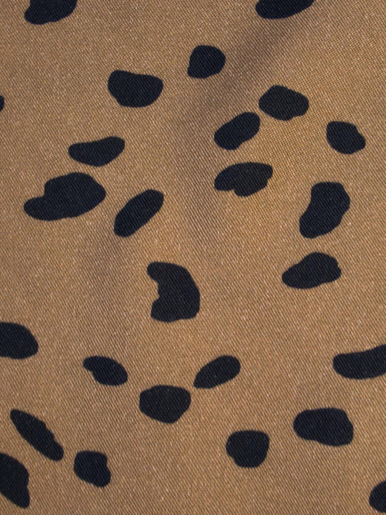 May include: Close-up of a tan fabric with a repeating pattern of dark navy blue irregular shapes. The shapes are scattered across the surface, creating a spotted or speckled effect. The fabric has a subtle texture.