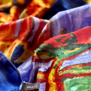 May include: Close-up of a colorful silk scarf with abstract patterns in shades of red, blue, yellow, and purple. The scarf has a tag that reads "100% Silk". The fabric is soft and flowing.