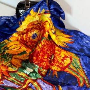 May include: A silk scarf featuring a vibrant sunflower design in shades of yellow, orange, red, and green against a deep blue background. The scarf is draped over a black handbag with a brown handle.