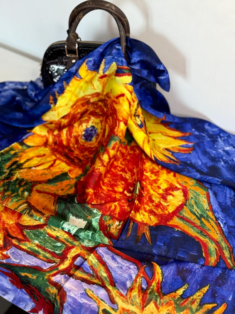 May include: A silk scarf featuring a vibrant sunflower design in shades of yellow, orange, red, and green against a deep blue background. The scarf is draped over a black handbag with a brown handle.