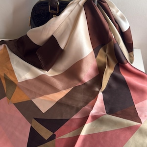 May include: A geometric patterned silk scarf in shades of brown, beige, and pink is draped over a brown leather handbag. The scarf features a repeating pattern of angular shapes and a glossy finish, adding a touch of elegance.