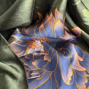 May include: A dark green silk scarf with a floral pattern in shades of blue and orange. The scarf is folded, revealing the intricate design. A small tag reads "100% SILK".