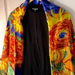 May include: A black crew neck shirt is displayed under a colorful, patterned shawl. The shawl features a vibrant design with yellow, red, blue, and green hues. The text "FASHION NOVA" is visible on the shirt.