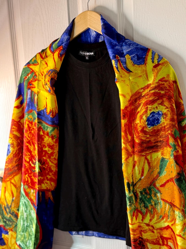 May include: A black crew neck shirt is displayed under a colorful, patterned shawl. The shawl features a vibrant design with yellow, red, blue, and green hues. The text "FASHION NOVA" is visible on the shirt.