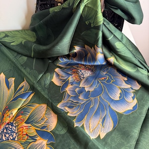 May include: A dark green silk scarf with a large floral pattern in blue and peach. The scarf is draped over a black handbag with brown leather handles. The floral design features detailed petals and leaves.