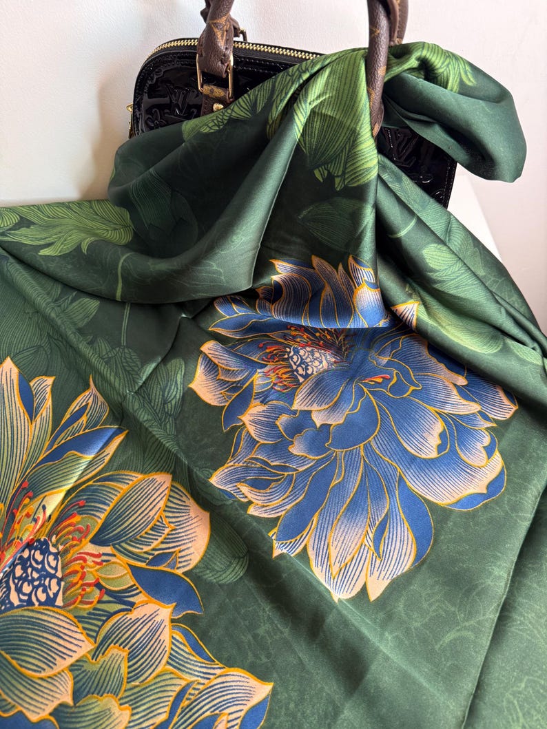 May include: A dark green silk scarf with a large floral pattern in blue and peach. The scarf is draped over a black handbag with brown leather handles. The floral design features detailed petals and leaves.