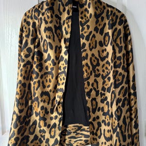 May include: A leopard print jacket with a black interior. The jacket is hanging on a wooden hanger against a white background. The leopard print features brown and black spots on a tan background.