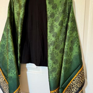 Può includere: Sciarpa verde con stampa floreale e bordi con stampa leopardata, drappeggiata su un top nero. La sciarpa ha uno sfondo verde scuro con motivi floreali neri. Il bordo presenta un design con stampa leopardata con dettagli arancioni e neri. Il marchio "FASHION NOVA" è visibile.