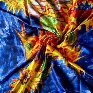 May include: A vibrant silk scarf with a painterly design. The scarf features large yellow sunflowers with red and orange accents against a deep blue background. The fabric is folded, creating a dynamic display of color and texture.