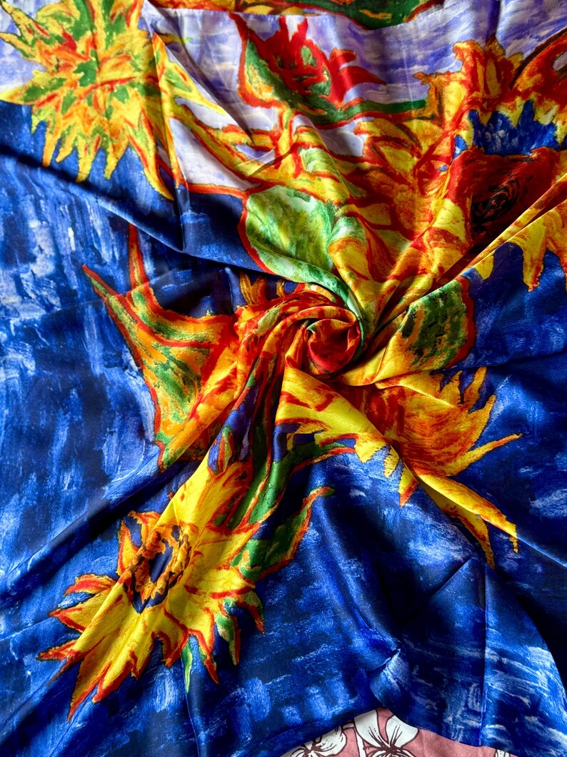 May include: A vibrant silk scarf with a painterly design. The scarf features large yellow sunflowers with red and orange accents against a deep blue background. The fabric is folded, creating a dynamic display of color and texture.