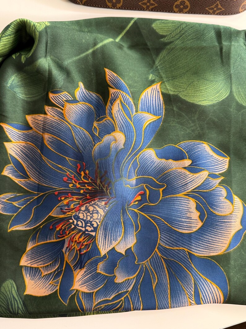 May include: A dark green silk scarf featuring a large blue and gold floral design. The flower has intricate details with a mix of blue, gold, and red accents. The background has green leaves.