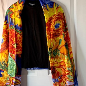 May include: A black top with a colorful scarf draped over it. The scarf features a vibrant floral design in yellow, red, blue, and green, reminiscent of a painting. The scarf is hanging on a wooden hanger.