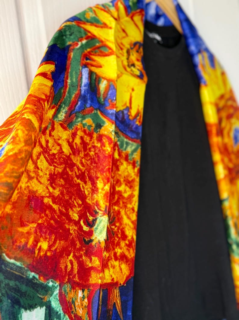May include: A vibrant, colorful kimono-style garment with a floral design in shades of yellow, red, blue, and green. The kimono is draped over a black top, creating a striking contrast. The garment is hanging on a wooden hanger.