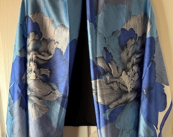 May include: A blue floral scarf with a large flower design in shades of blue, gray, and white. The scarf is made of a silky material and is hanging on a hanger. The scarf has a white border.
