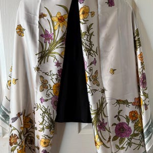 May include: White silk scarf with a floral design, including yellow, purple, and orange flowers, butterflies, and insects. The scarf has a green and blue border and is draped over a black garment.