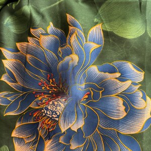 May include: A dark green scarf with a large blue and yellow floral design. The flower has intricate details with a mix of blue, yellow, and orange hues. The background features green leaves, creating a natural and elegant aesthetic.