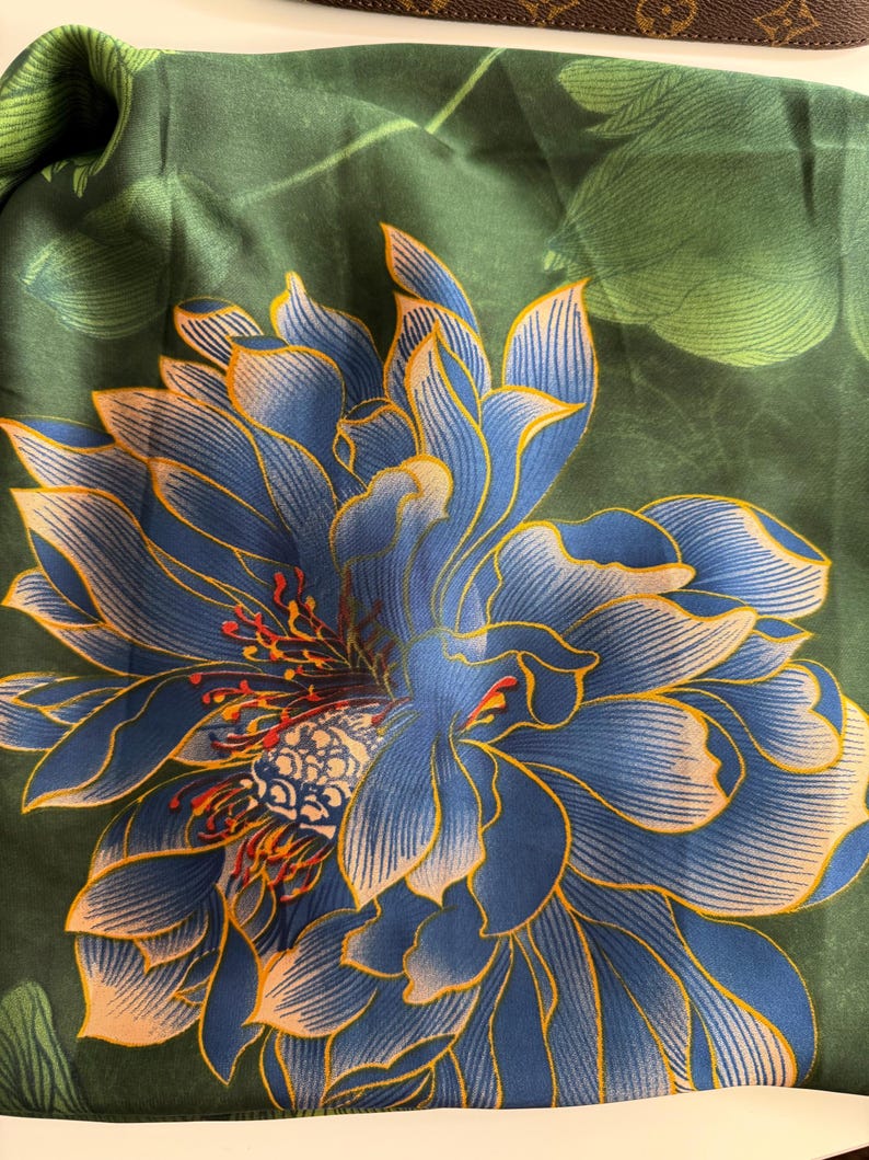 May include: A dark green scarf with a large blue and yellow floral design. The flower has intricate details with a mix of blue, yellow, and orange hues. The background features green leaves, creating a natural and elegant aesthetic.