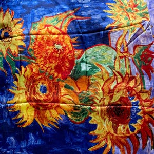 A scarf featuring a vibrant floral design reminiscent of Van Gogh's 'Sunflowers.' The scarf showcases large sunflowers with yellow petals and dark centers against a deep blue background. The artwork style includes visible brushstrokes and a textured appearance, with hints of green leaves and orange accents.