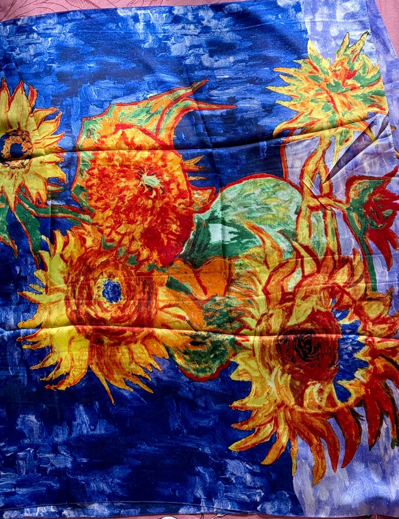 A scarf featuring a vibrant floral design reminiscent of Van Gogh's 'Sunflowers.' The scarf showcases large sunflowers with yellow petals and dark centers against a deep blue background. The artwork style includes visible brushstrokes and a textured appearance, with hints of green leaves and orange accents.
