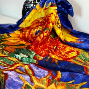 silk scarf with a vibrant sunflower design. The scarf features a blue background with large yellow, orange, and red sunflower blooms. The scarf is draped over a black handbag, showcasing the colorful floral pattern.