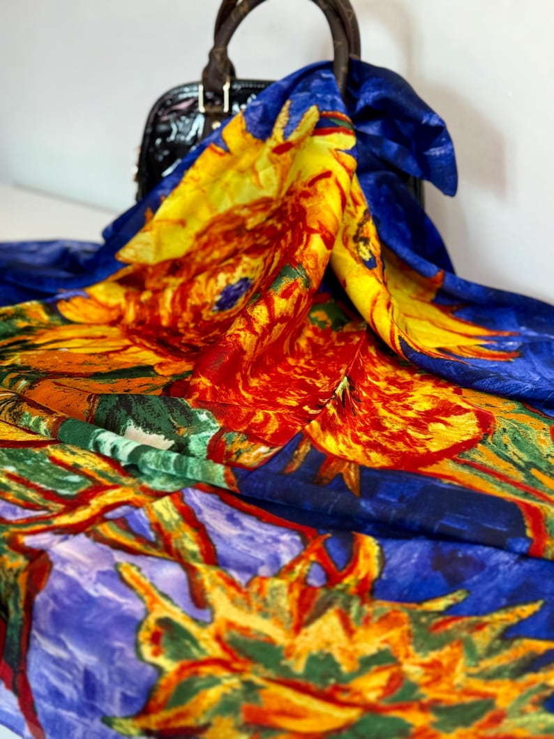 silk scarf with a vibrant sunflower design. The scarf features a blue background with large yellow, orange, and red sunflower blooms. The scarf is draped over a black handbag, showcasing the colorful floral pattern.