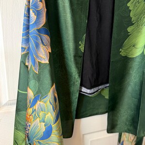 May include: A green floral kimono with blue and peach-colored flower designs. The kimono is made of a silky material and has long sleeves. The background is a white door.