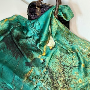 May include: A teal and green silk scarf with a nature-inspired design featuring trees, birds, and floral elements. The scarf is draped over a black handbag with brown leather handles.