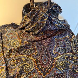 May include: A black handbag with a wooden handle is paired with a paisley patterned scarf. The scarf features a complex design in shades of blue, yellow, and red on a dark background. The scarf is draped over the handbag.