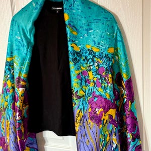 May include: A vibrant, floral-patterned scarf in shades of turquoise, purple, and yellow, draped over a black top. The scarf features a botanical design with the text "FASHIONOVA" on the label. The scarf is hanging on a wooden hanger.
