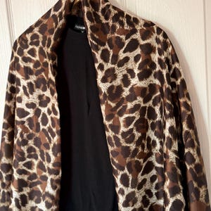 Puede incluir: Un chal con estampado de leopardo drapeado sobre una parte superior negra. El chal presenta un patrón de manchas marrones y beige. La palabra "FASHION" es visible en la etiqueta del chal. La prenda está colgada en una percha.