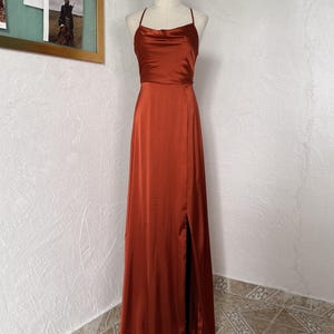 Puede incluir: Un vestido de satén color óxido con cuello de cisne y tirantes finos. El vestido largo hasta el suelo presenta una abertura alta en el lado izquierdo. El vestido se exhibe en un maniquí.