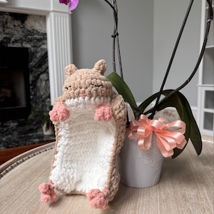 May include: A crocheted plush toy shaped like a small animal with light brown fur, a white belly, and pink paws. The creature has closed eyes and a small pink nose. The texture is soft and yarn-based, featuring a rounded head and small ears.