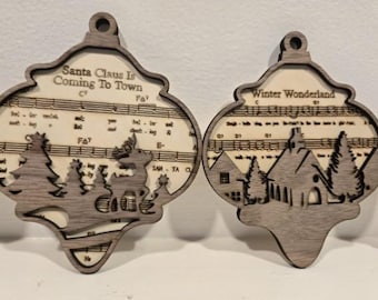 May include: Two wooden Christmas ornaments. One features a deer and trees with the text "Santa Claus Is Coming To Town" over sheet music. The other has a church and trees with the text "Winter Wonderland" over sheet music.