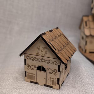 May include: A miniature wooden house with a gabled roof and decorative cutouts. The number "15" is displayed above the arched doorway. The house is made of light-coloured wood with a darker brown roof, and is likely a decorative item.